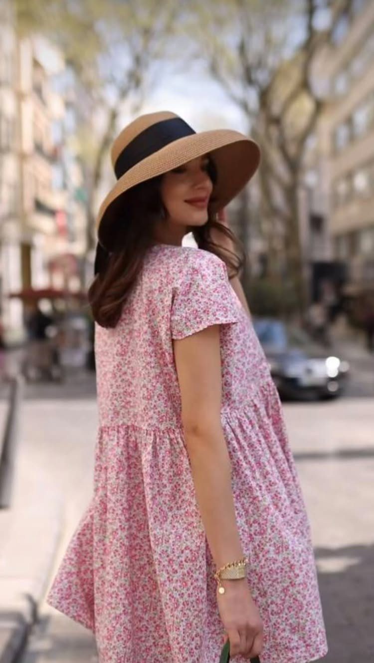 A woman in a pink floral smock dress and straw hat standing on a city street.