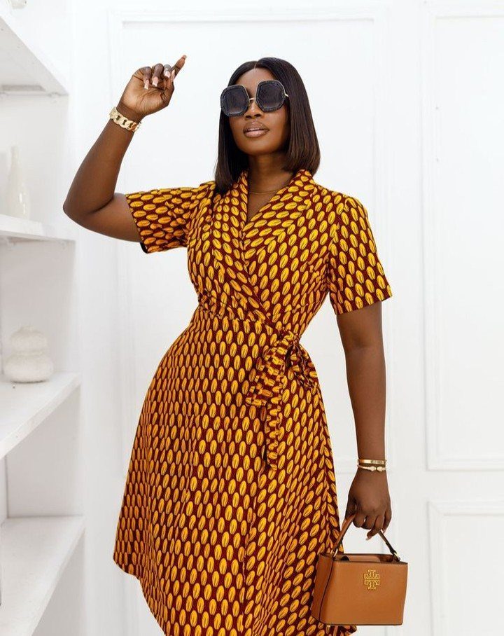 A woman posing in a patterned mustard yellow wrap dress with a waist tie.