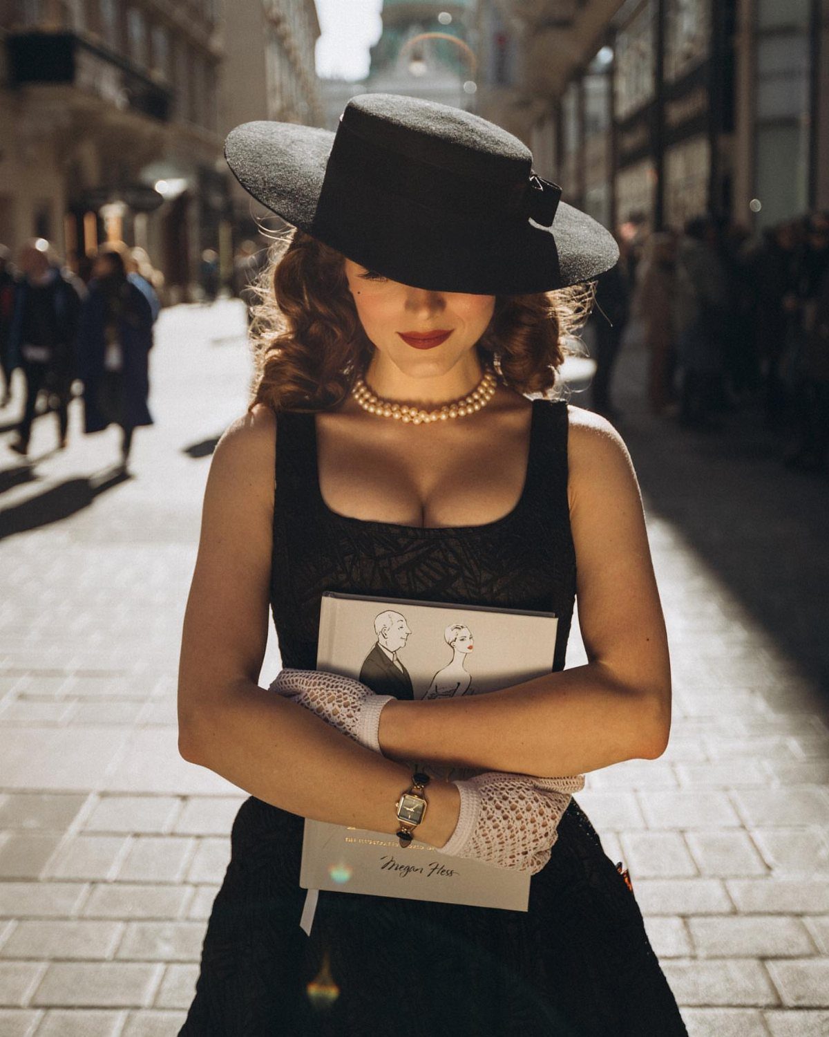 A woman wearing a black vintage dress, wide-brimmed hat, pearls, and lace gloves outdoors.
