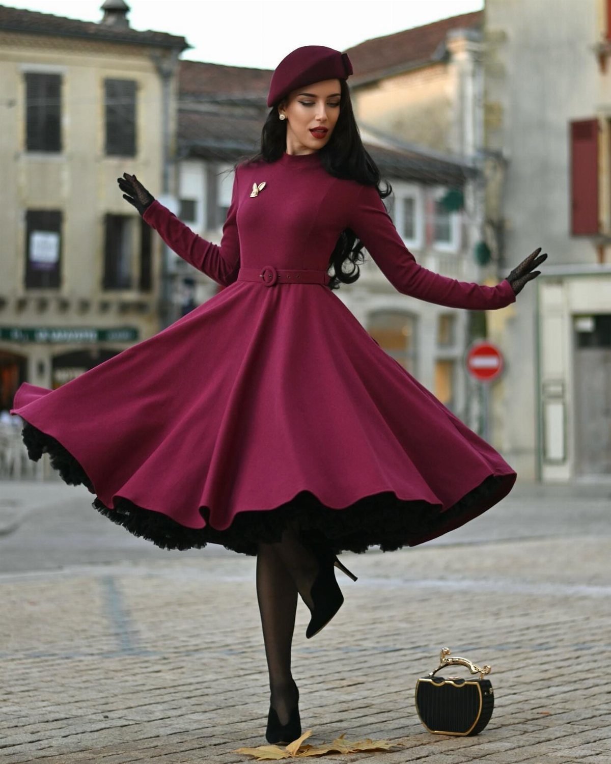 A woman in a burgundy vintage dress and beret posing in a cobblestone town square.
