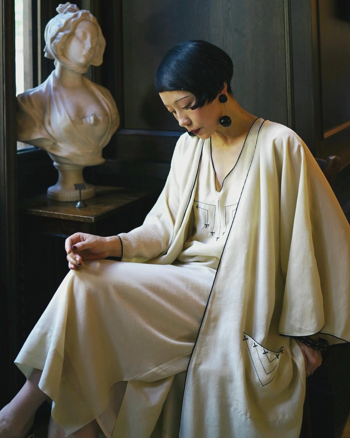 A woman in a cream vintage two-piece outfit sitting beside a white marble bust.