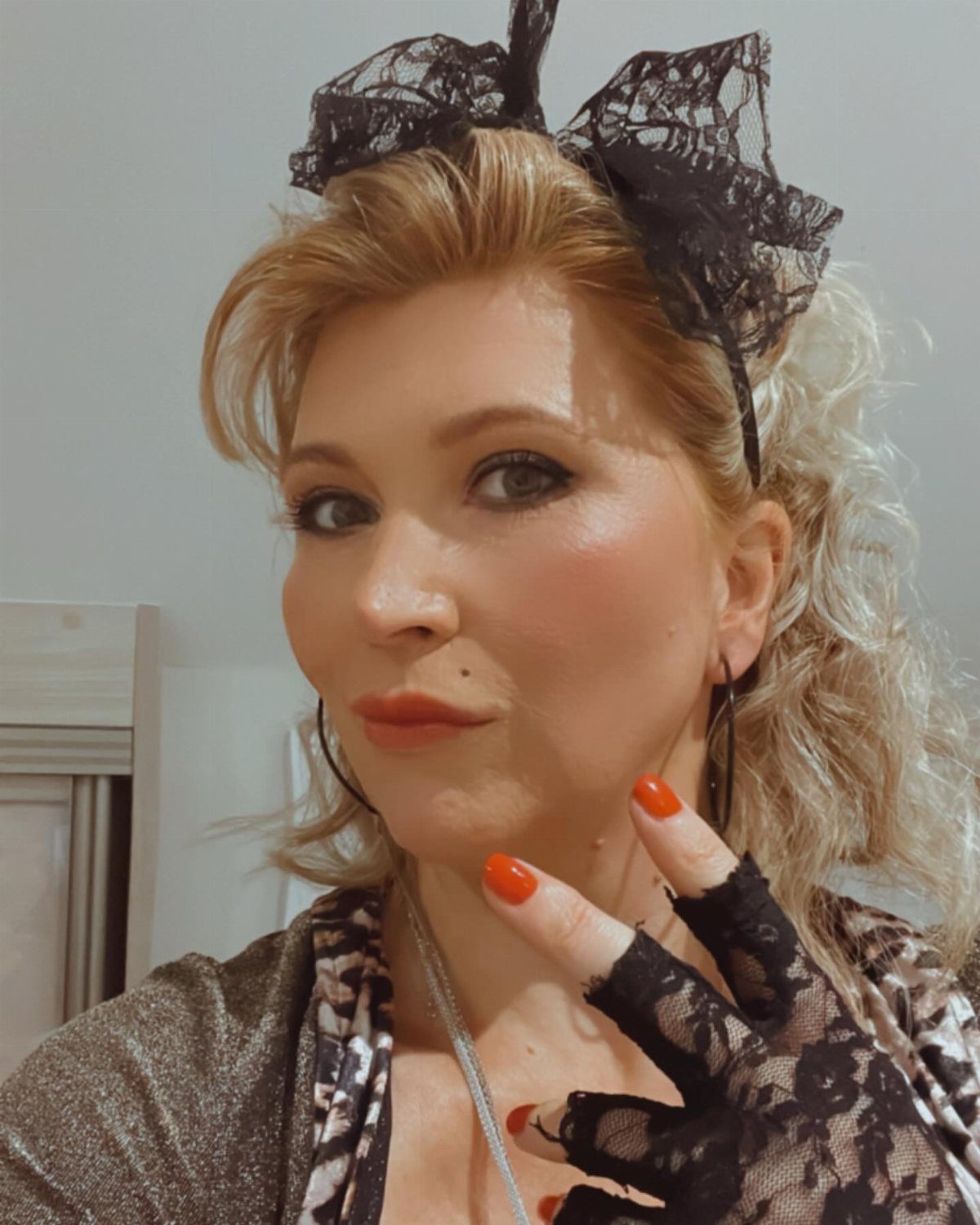 Blonde woman wearing black lace fingerless gloves and a large lace hair bow posing for a selfie.