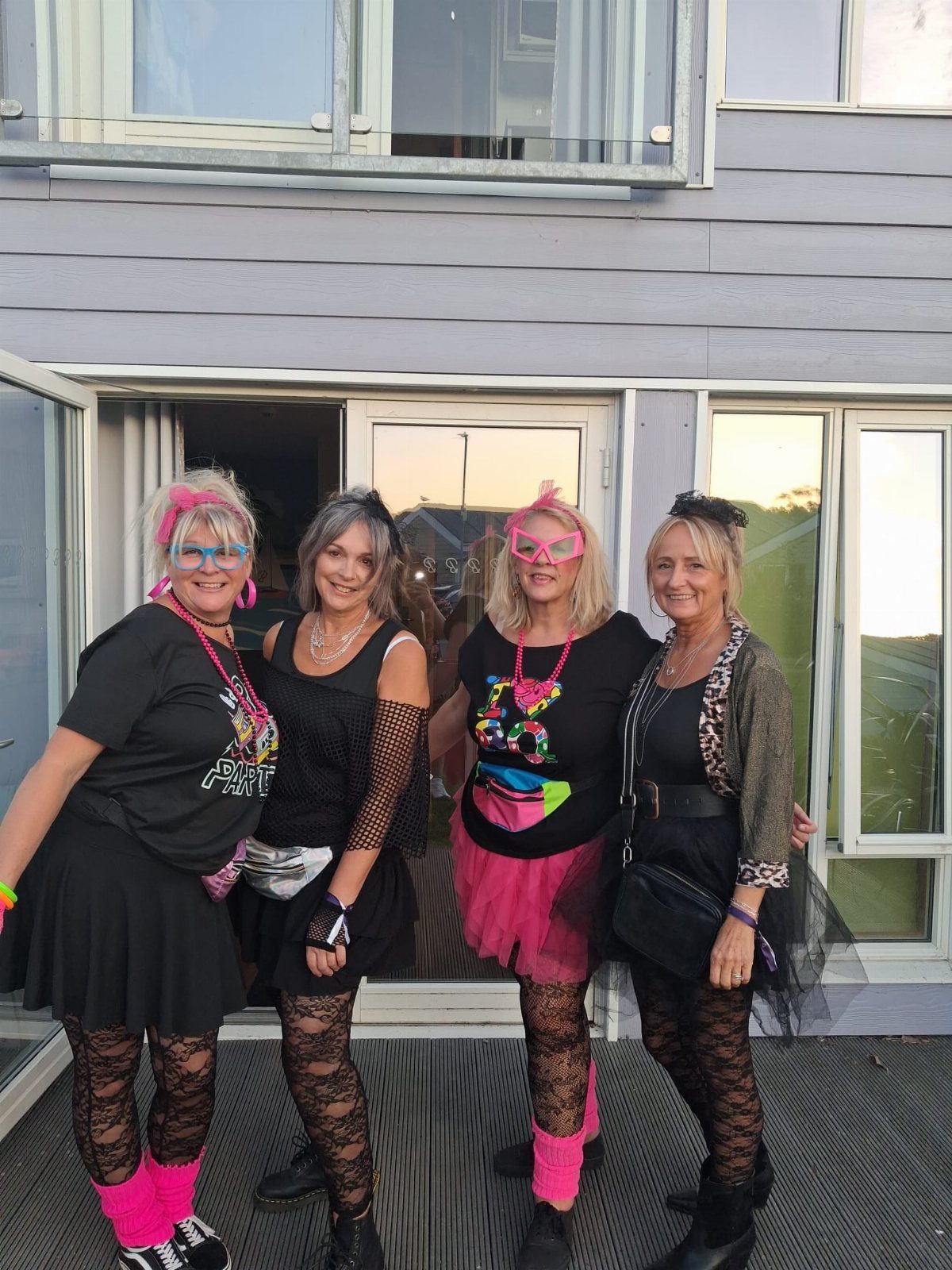 Four women dressed in 80s inspired costumes posing together in front of a house.