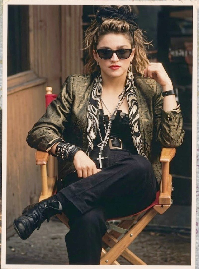 Madonna sitting in a chair wearing an olive jacket, sunglasses, and several layered necklaces with a cross.