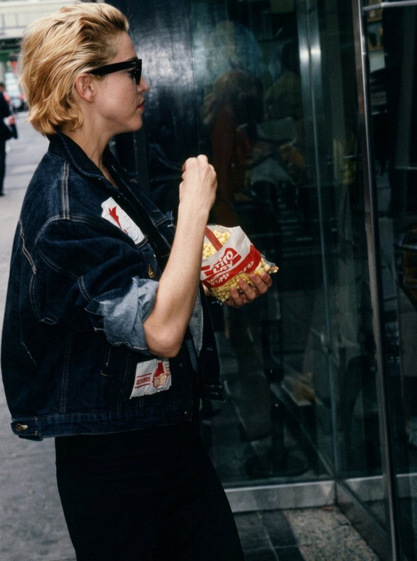 A blonde woman wearing an oversized denim jacket and sunglasses while holding a snack bag.