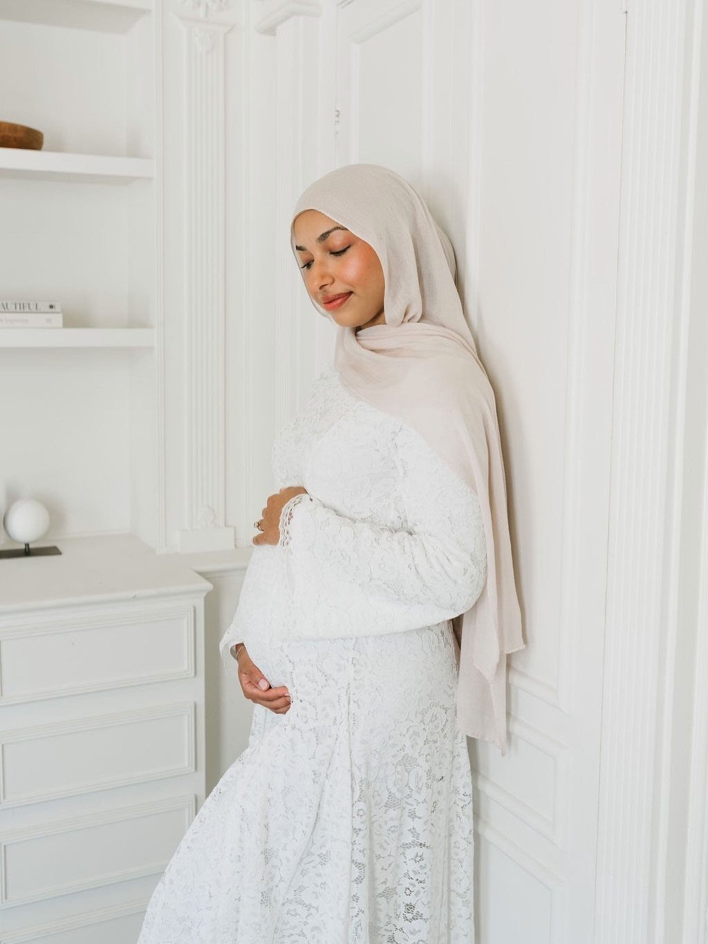Pregnant woman in a white lace maternity dress and beige hijab posing against a white wall.