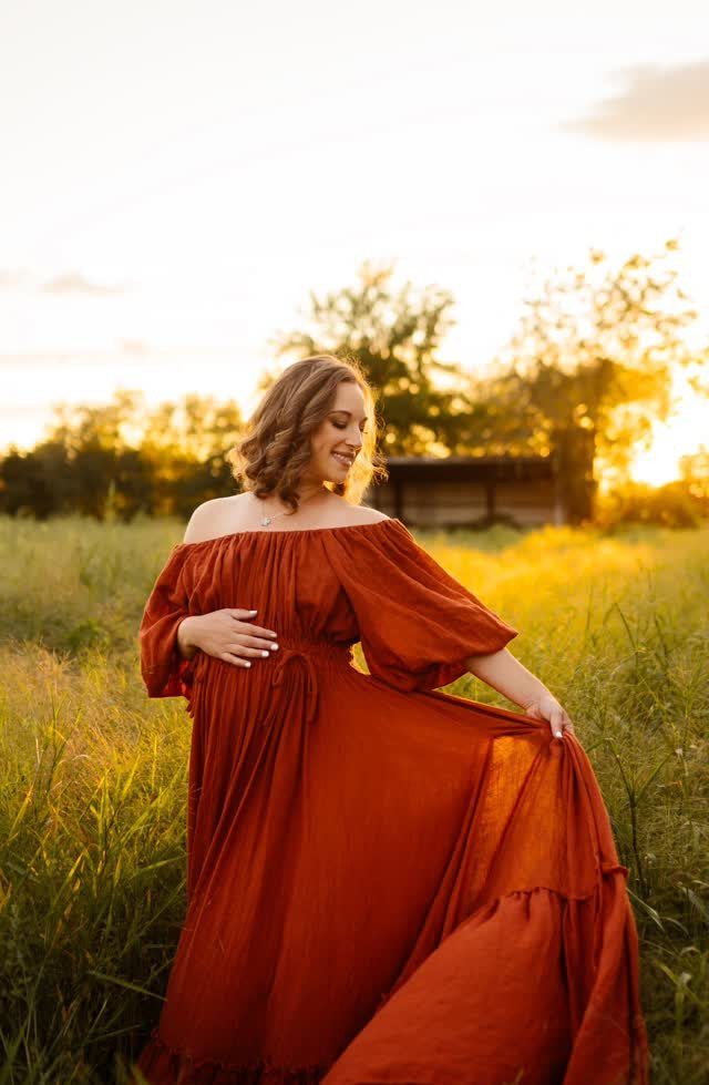 Pregnant woman in a rust-colored off-the-shoulder maxi dress standing in a sunlit grassy field.