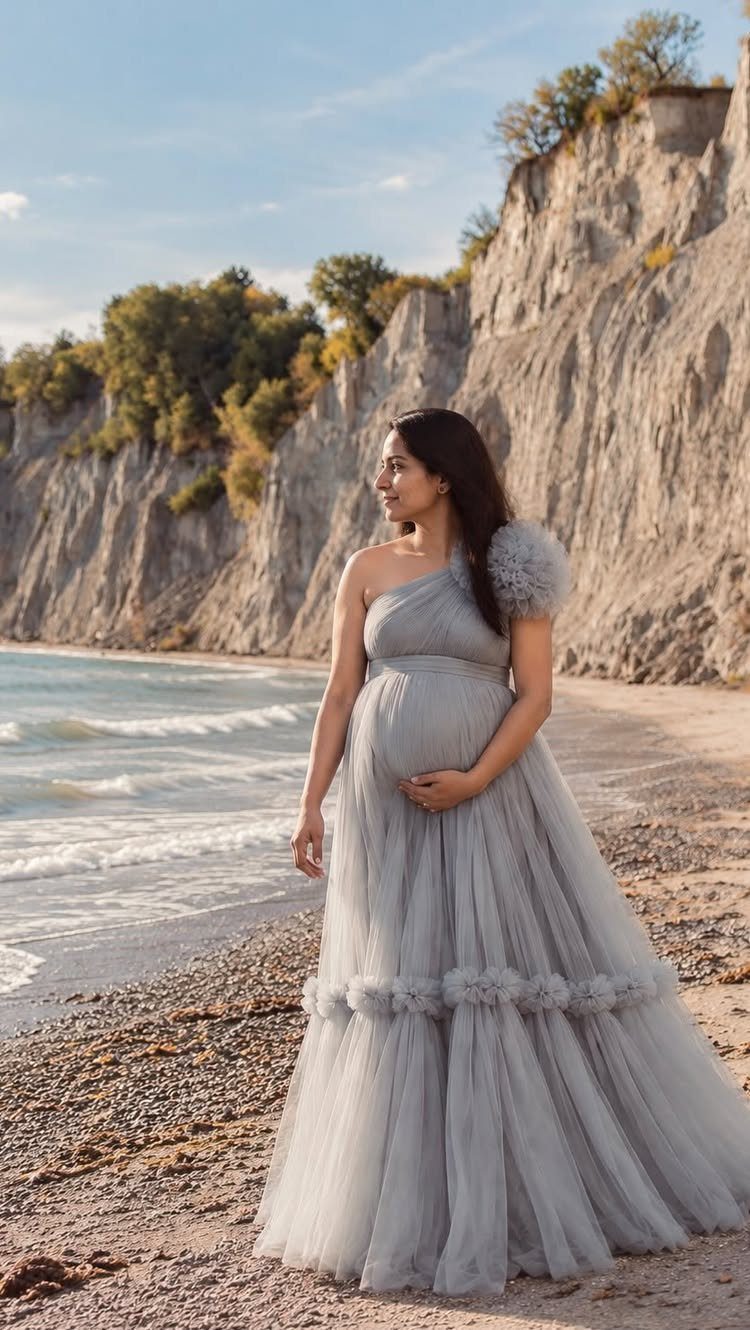 A pregnant woman in a flowing grey tulle gown standing on a beach near cliffs.