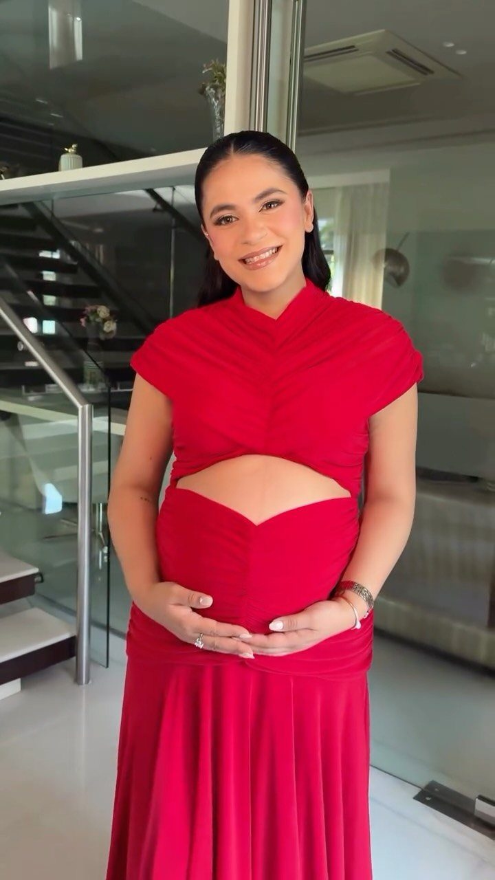 A smiling pregnant woman posing in a bright red two-piece maternity outfit indoors.