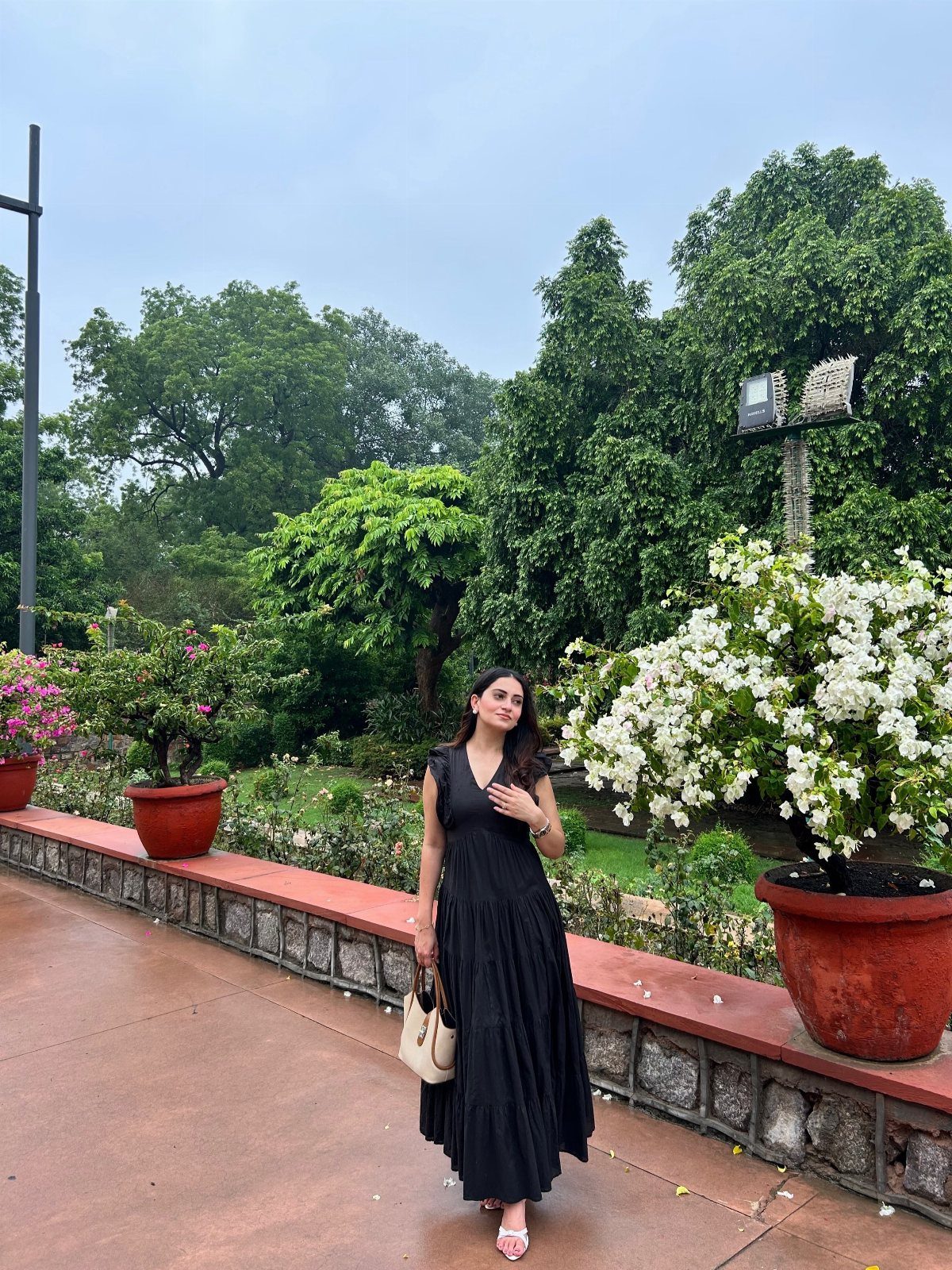 A woman posing in a black sleeveless maxi dress while holding a small beige handbag outdoors.
