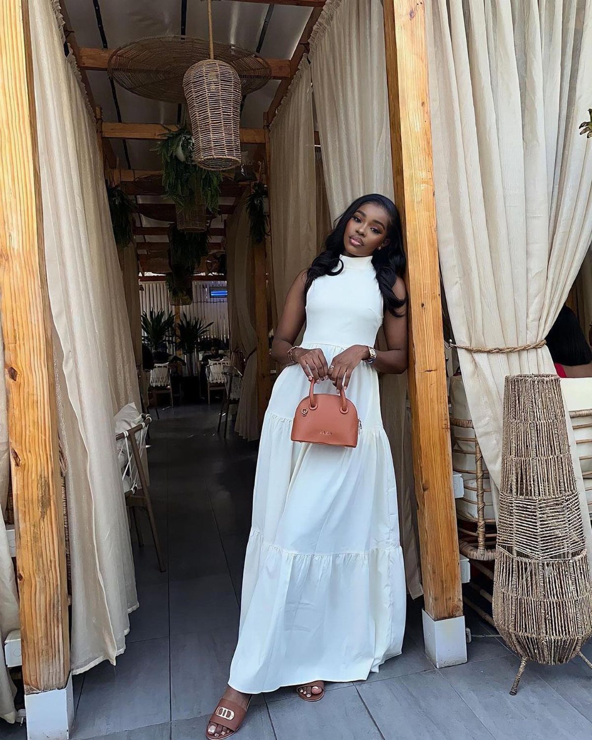 A woman in a sleeveless white maxi dress posing with a tan handbag under a pergola.