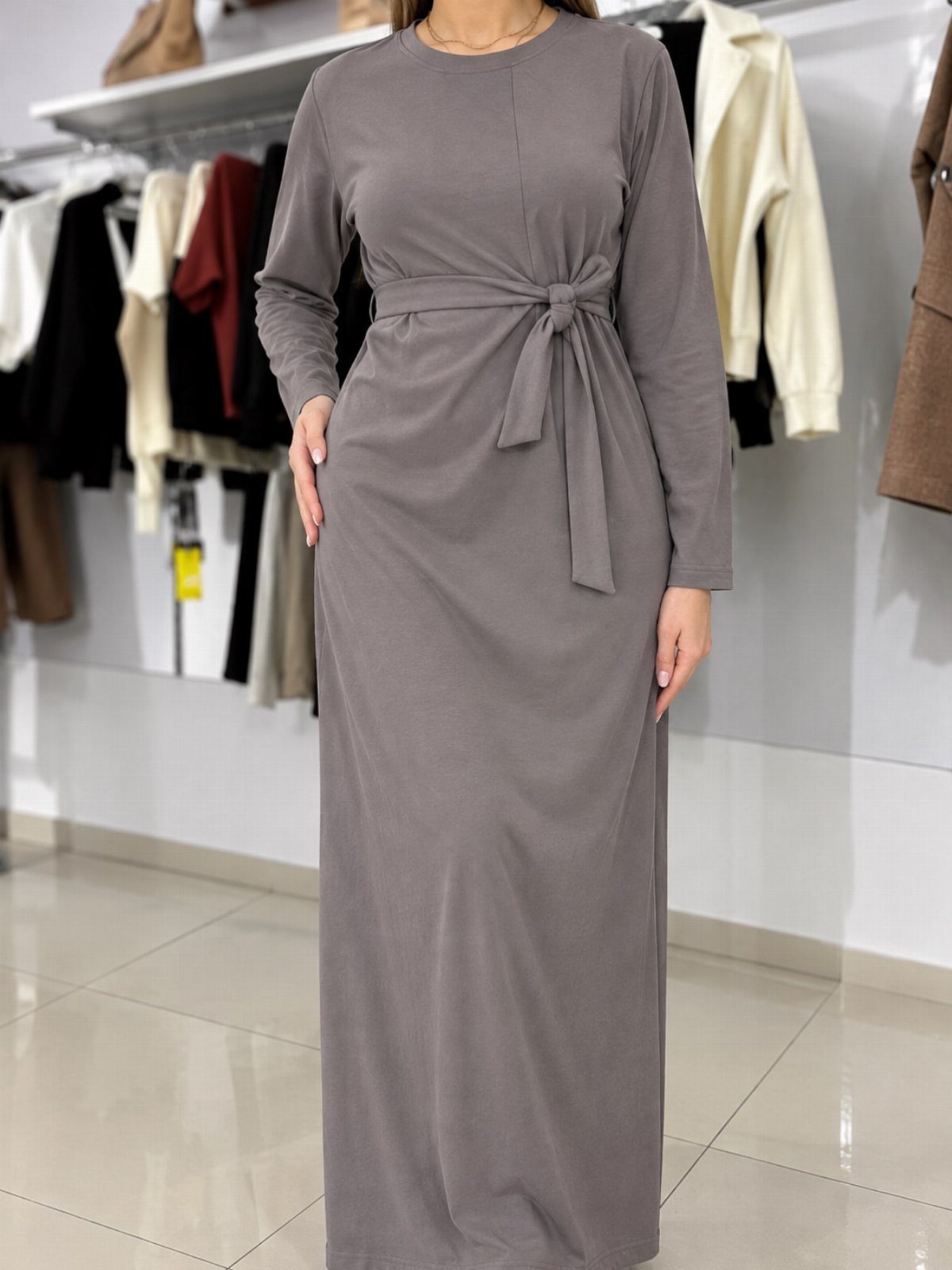 A person wearing a long-sleeved grey jersey maxi dress with a waist tie in a clothing store.