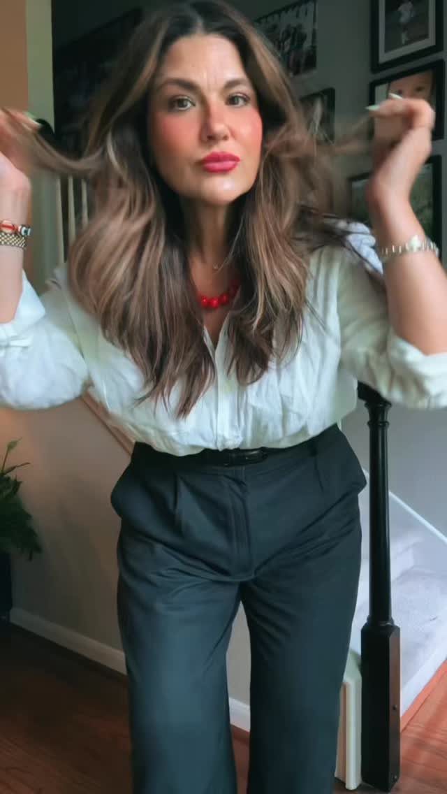 A woman posing in a white blouse and dark grey trousers with a red necklace.
