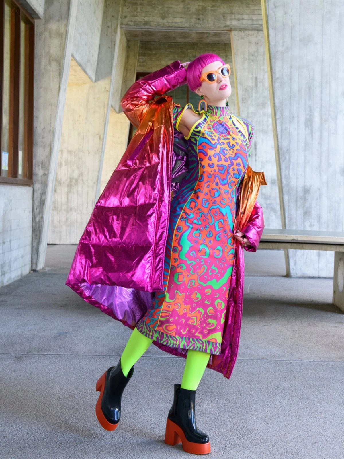 A person wearing a colorful patterned dress, magenta puffer coat, and lime green tights posing outdoors.