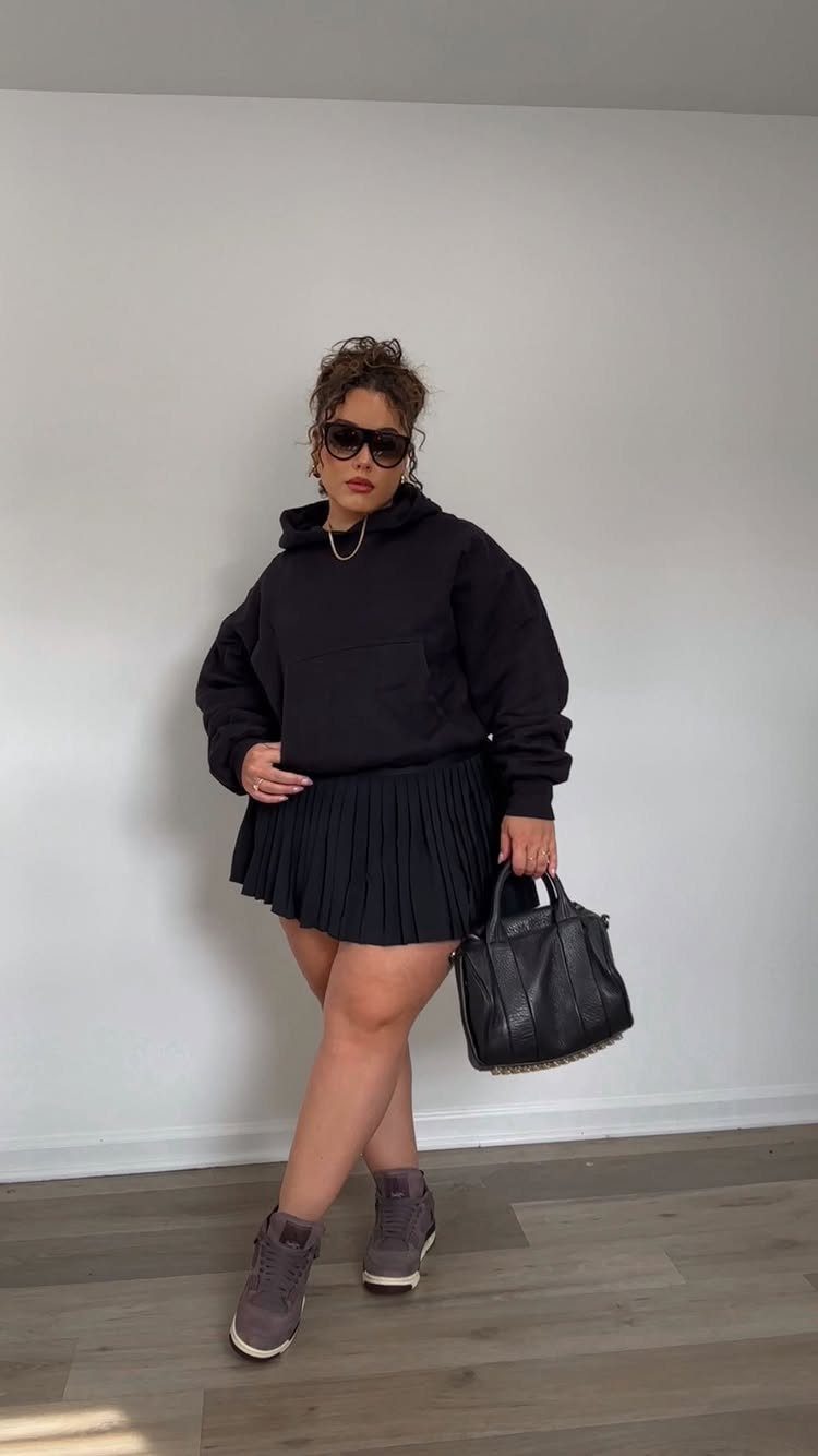 A woman wearing a black hoodie, pleated skirt, sunglasses, and grey sneakers posing indoors.