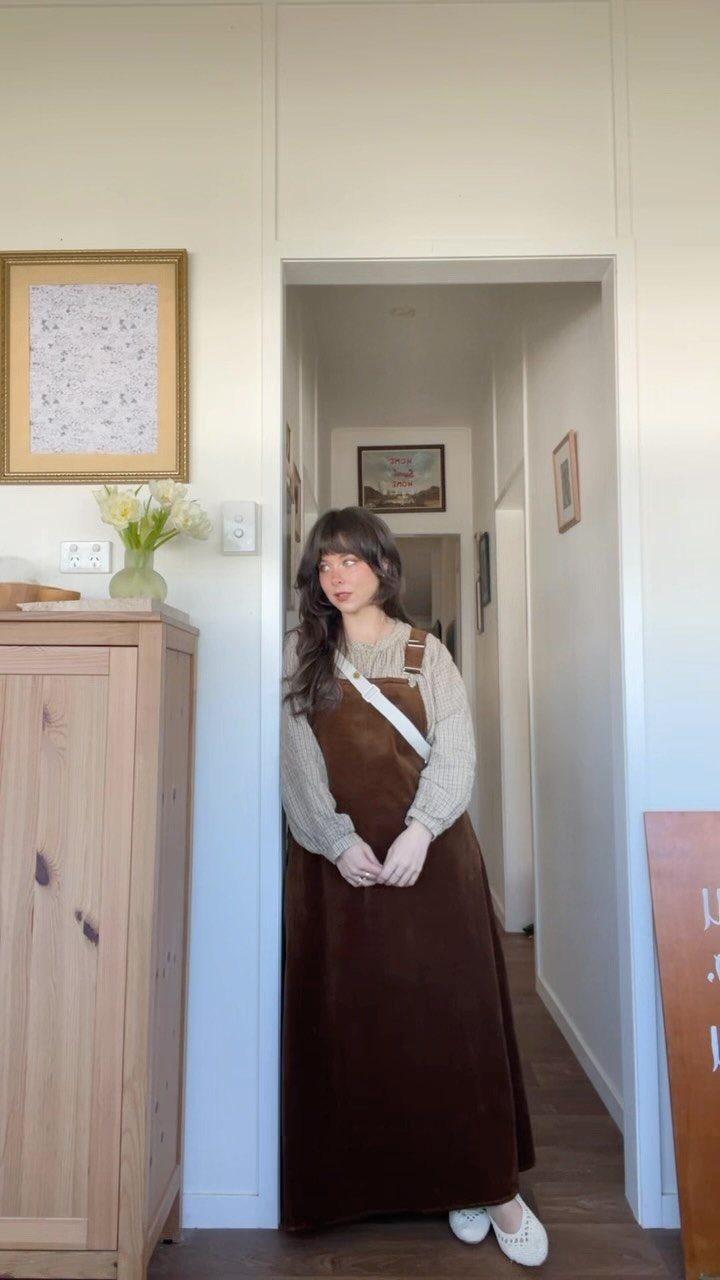 A woman wearing a long brown pinafore dress and a light blouse standing in a doorway.
