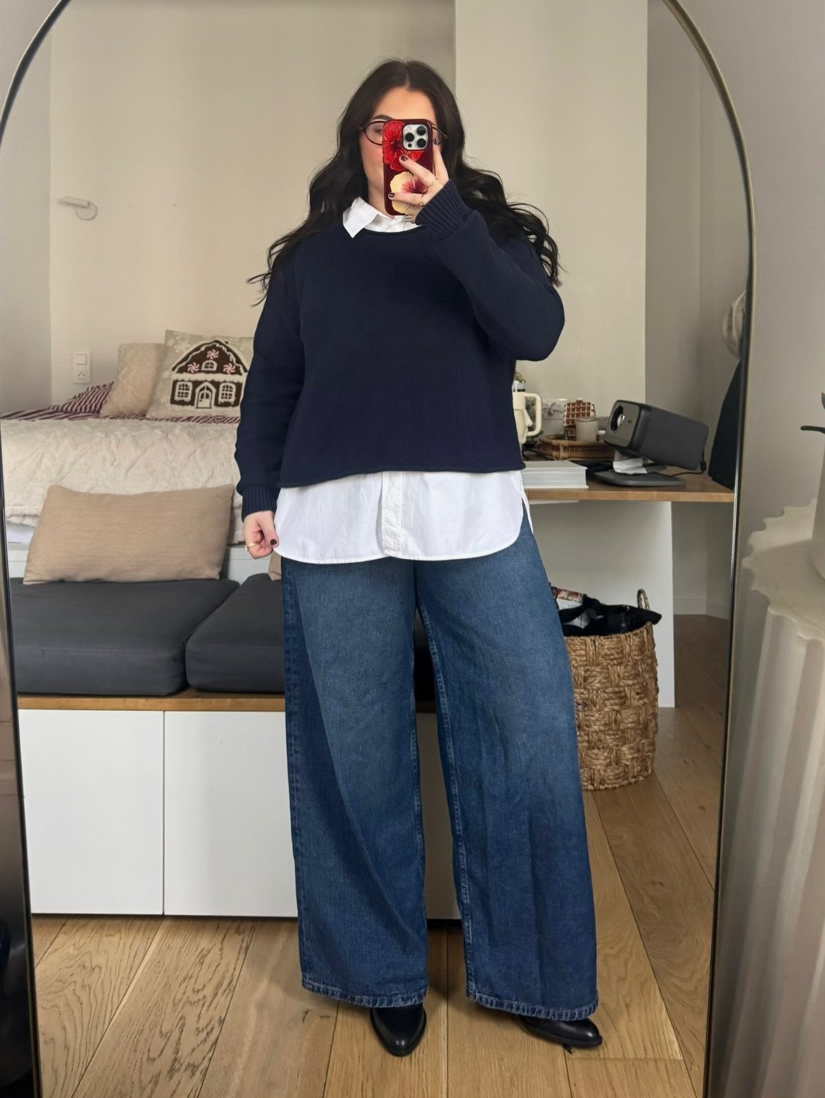 Woman in navy sweater, white shirt, and wide-leg blue jeans taking a mirror selfie.
