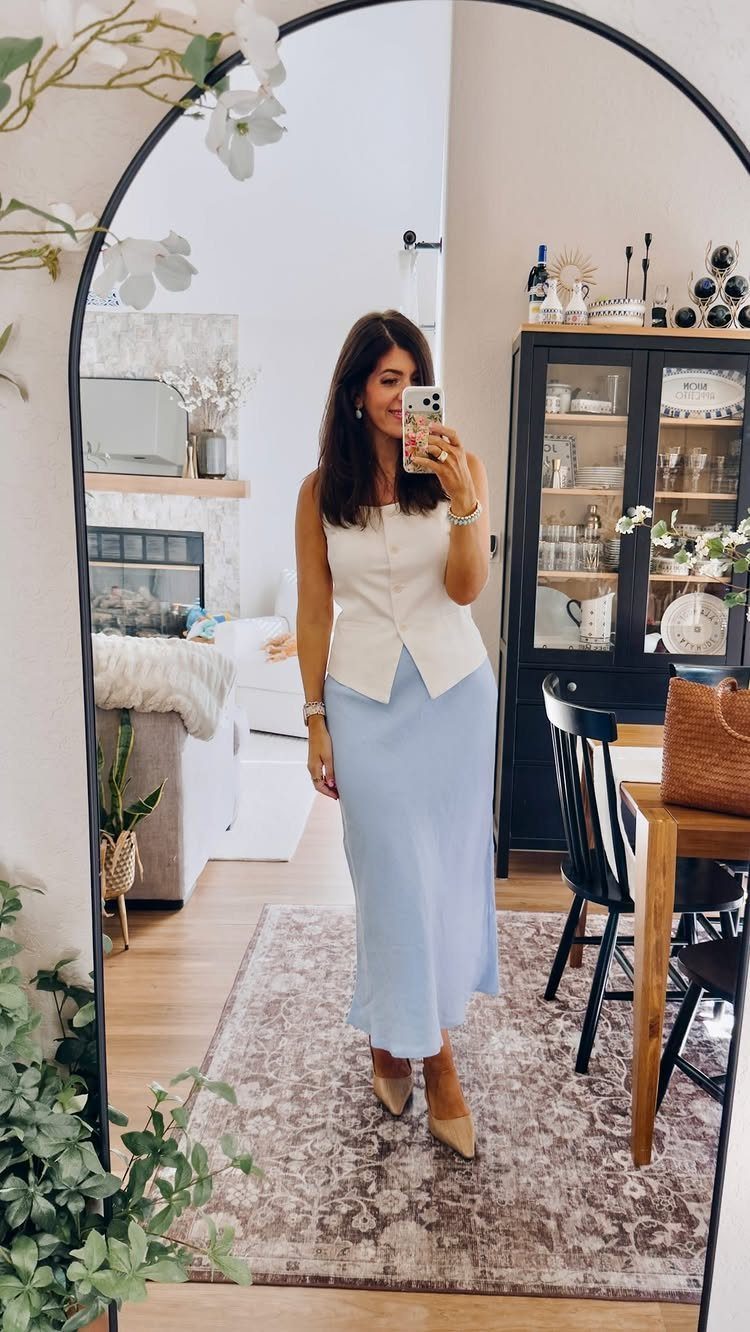 A woman posing in a mirror wearing a cream vest and light blue midi skirt.