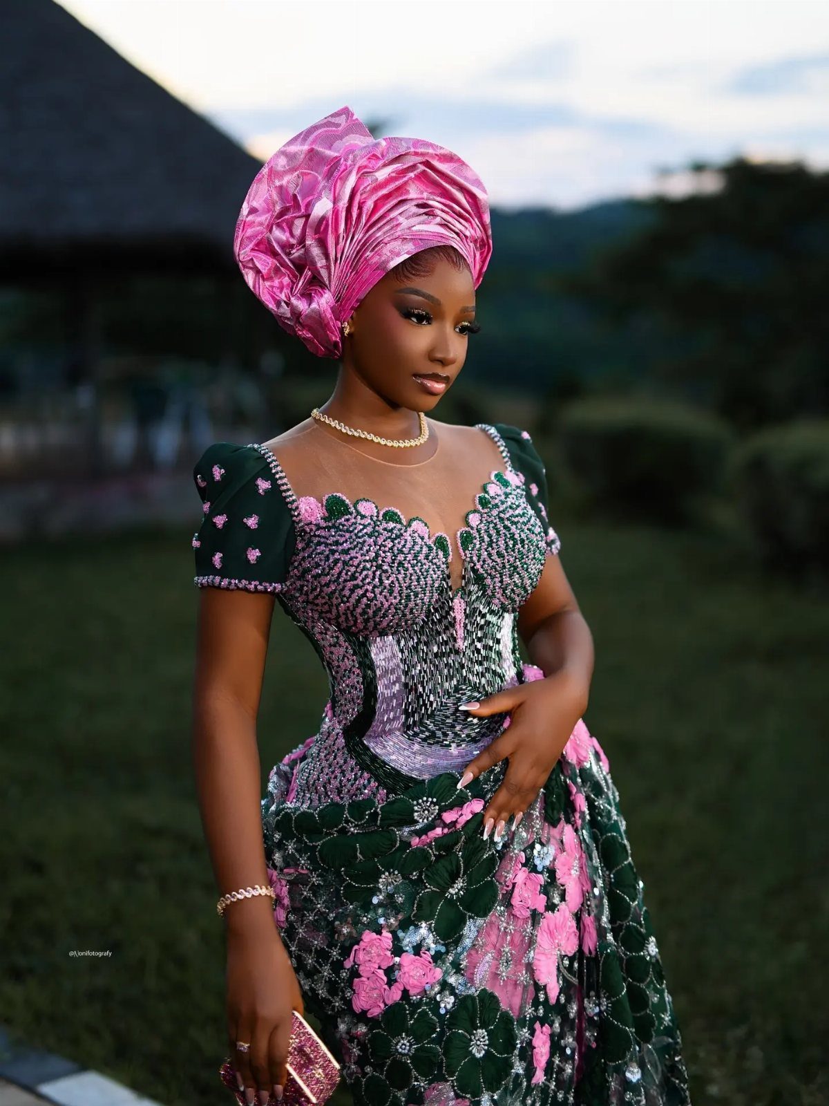 A woman wearing a pink gele and a green lace gown posing in a garden.