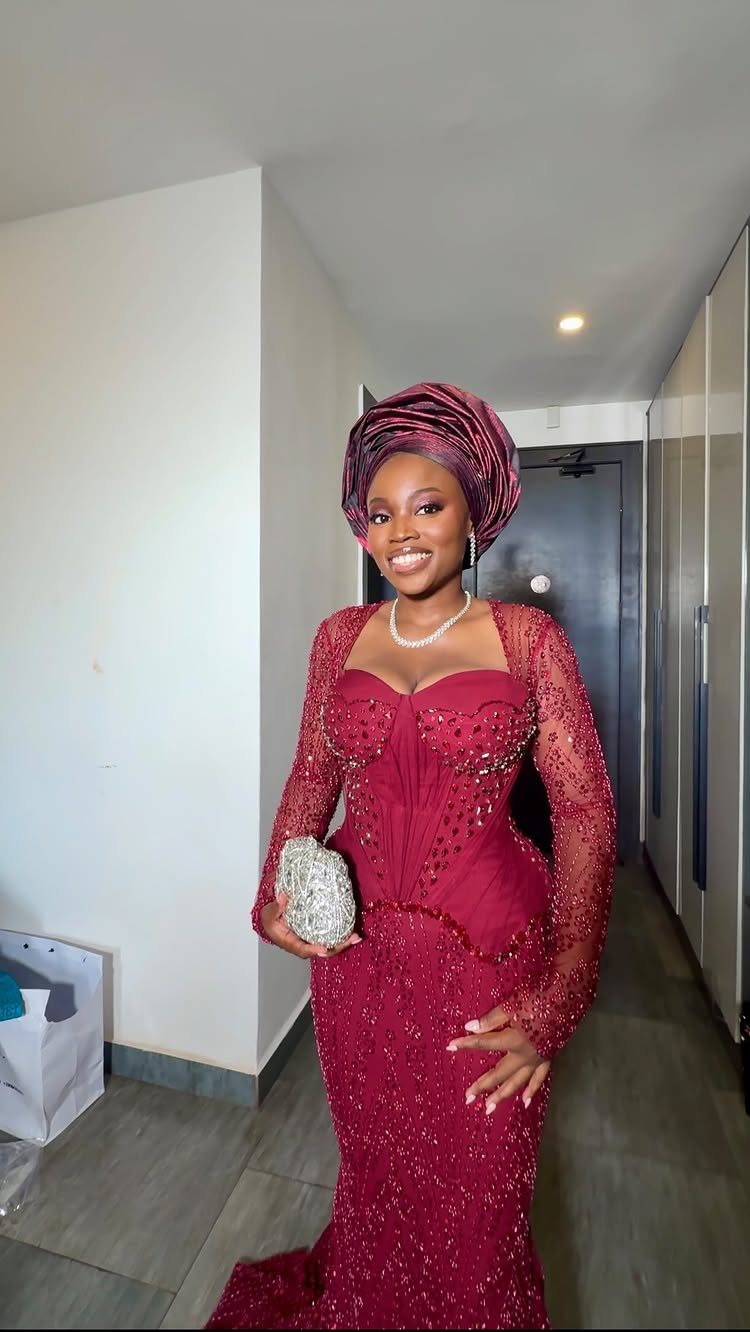 A woman in a beaded maroon lace gown and matching gele headtie smiling in a hallway.