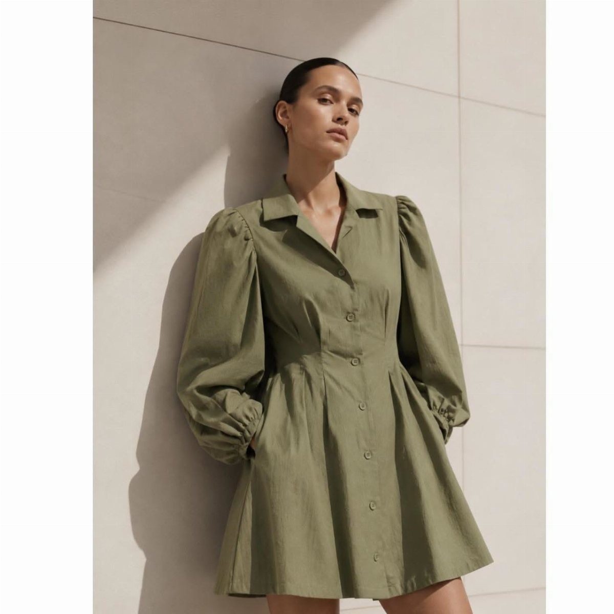 Woman posing in an olive green button-front mini dress with long puff sleeves against a wall.