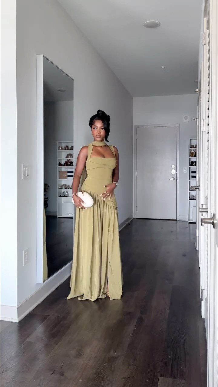 A woman posing in a long olive green halter maxi dress while holding a white clutch.