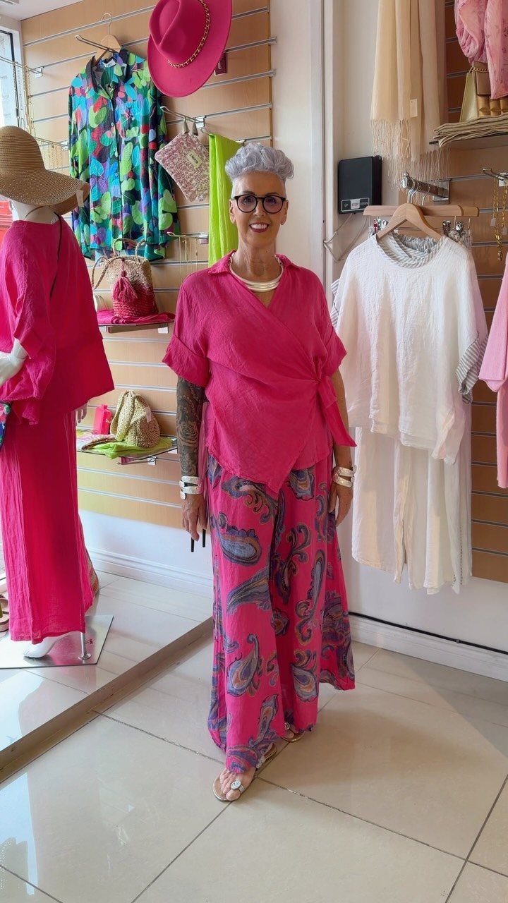 A smiling woman with grey hair wearing a bright pink wrap top and patterned wide-leg trousers.