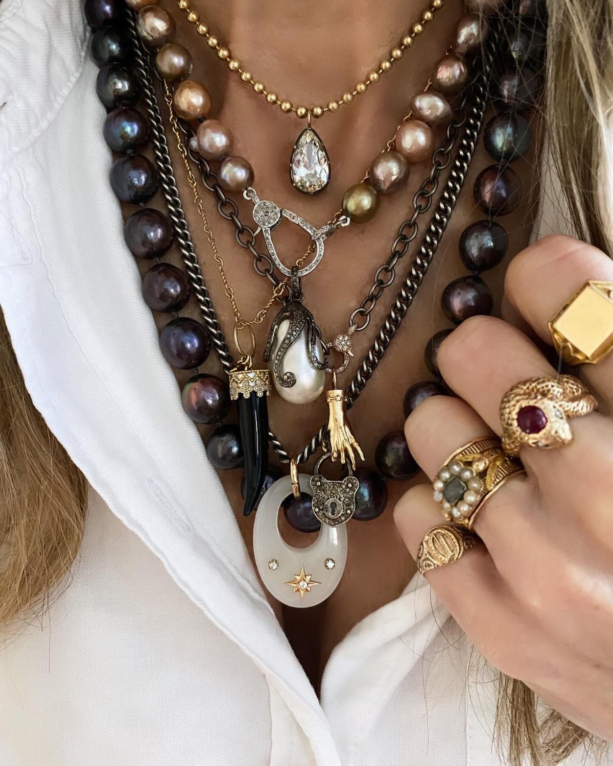 Layered pearl and gold necklaces with various pendants worn by a person with gold rings.