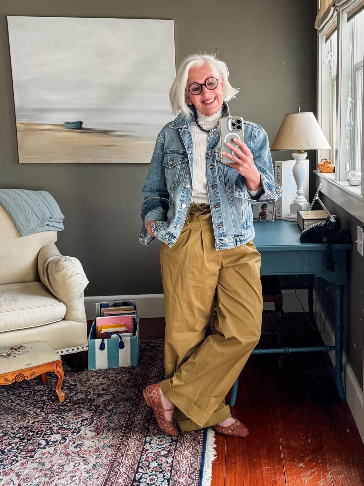 Smiling woman with white hair posing in a denim jacket and mustard yellow wide-leg trousers.