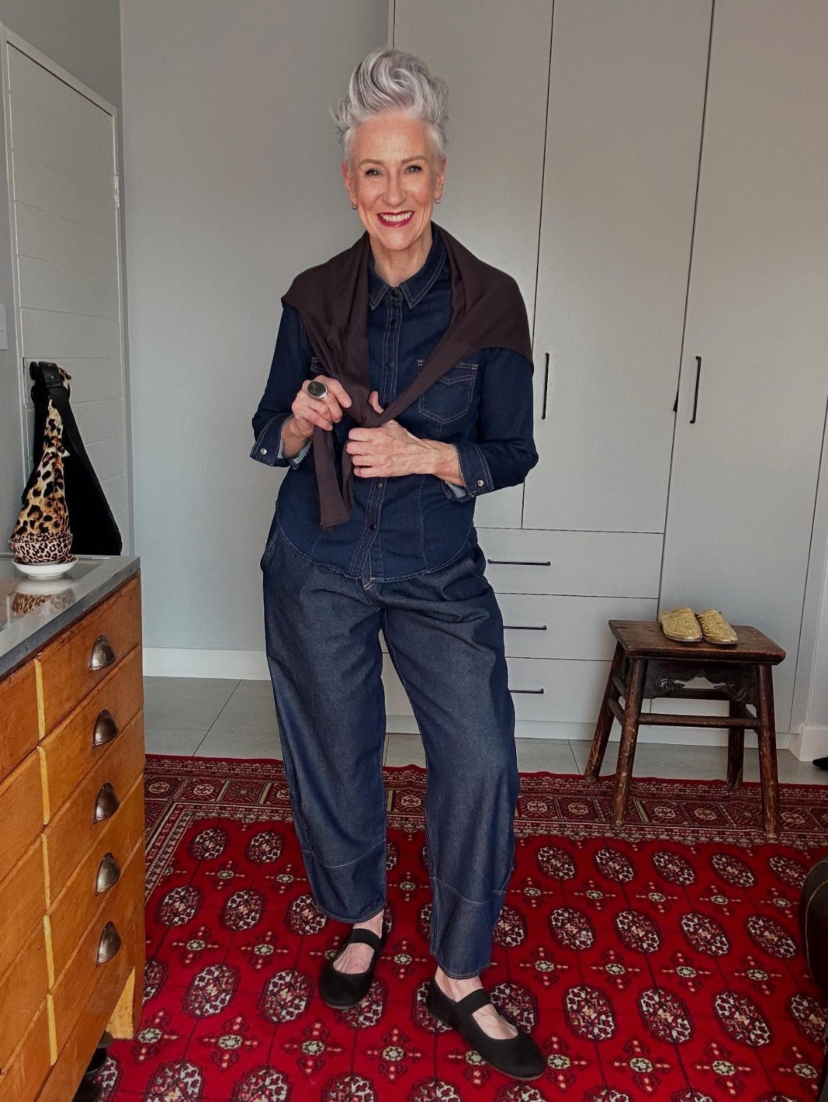 A smiling woman with grey hair wearing a denim shirt and loose denim trousers.