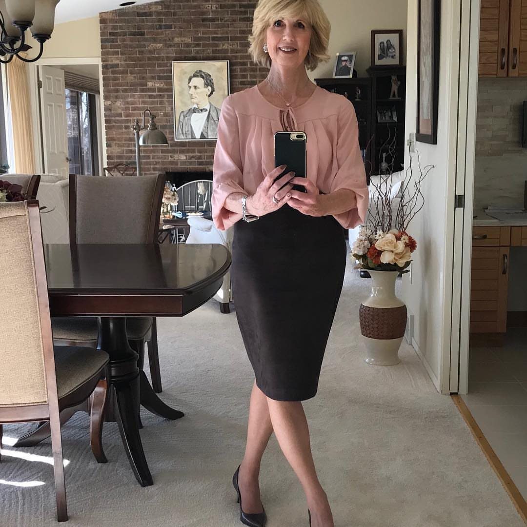 A woman in a pink blouse and dark skirt posing for a mirror selfie indoors.