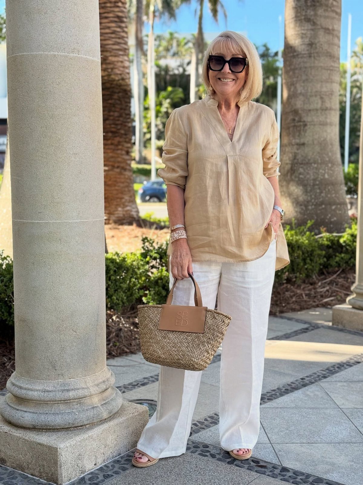 A smiling woman wearing a beige linen tunic and white relaxed linen trousers standing outdoors.