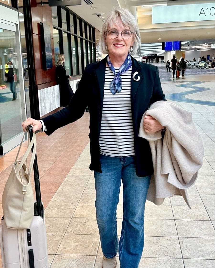 Smiling woman in a black blazer, striped shirt, and blue jeans walking through an airport terminal.