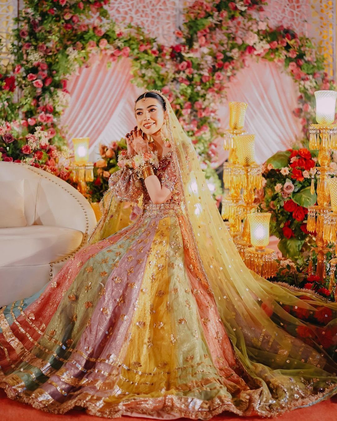 A smiling bride in a colorful embroidered lehenga sitting on a white sofa amidst flowers.