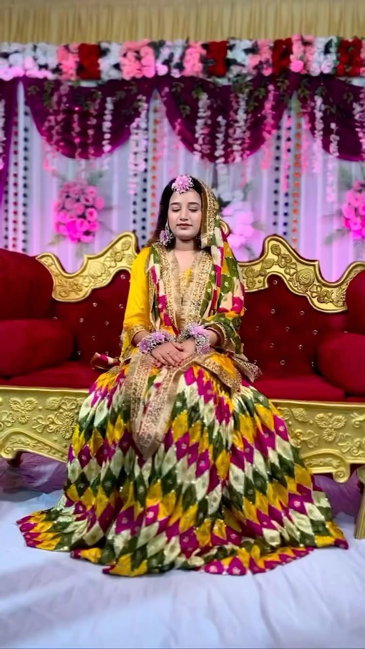 Woman sitting on a red sofa wearing a colorful yellow and patterned Pakistani gharara outfit.