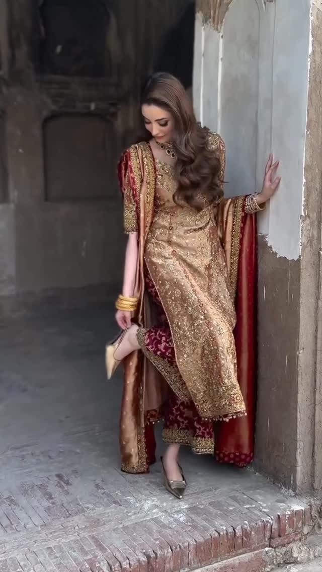 Woman wearing a gold embroidered long silk kameez and maroon patterned trousers against a stone wall.