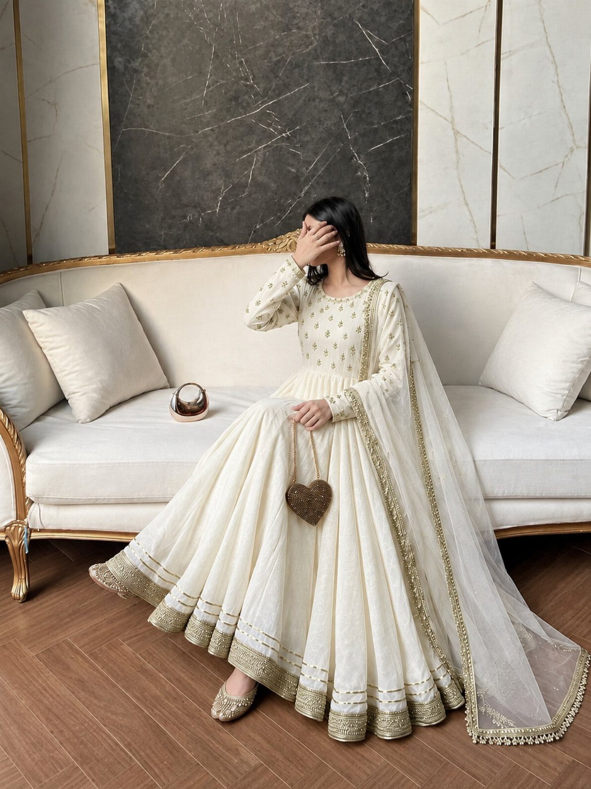 Woman in a white embroidered Anarkali frock sitting on a white sofa holding a gold bag.