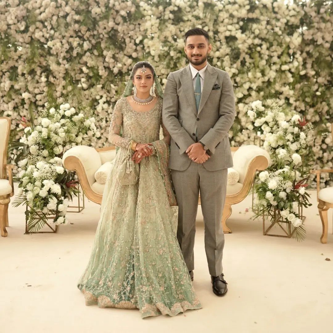 A bride wearing a sage green embroidered lehenga stands beside a man in a grey suit.