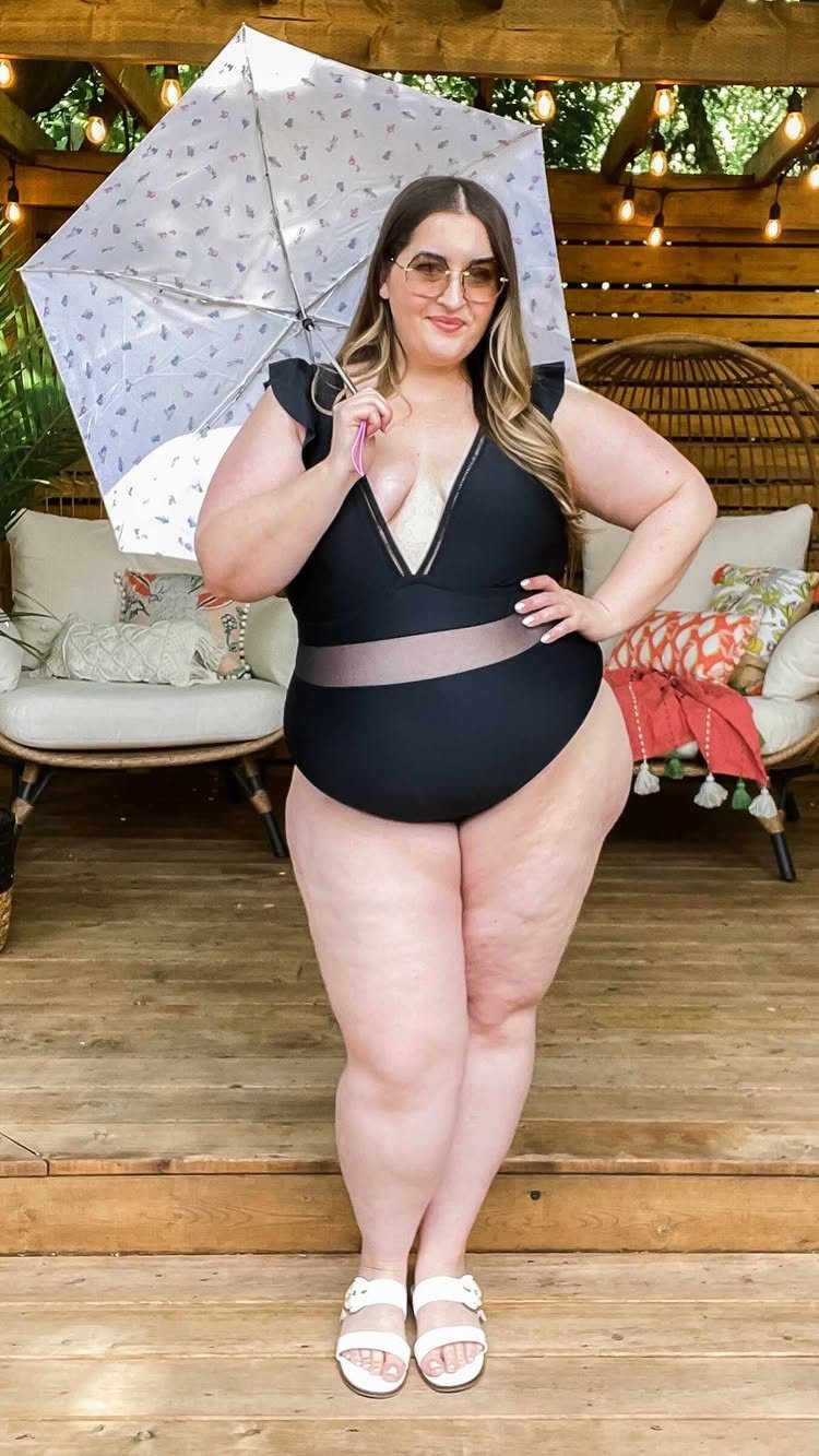 A plus size woman in a black one-piece swimsuit posing with a patterned umbrella outdoors.