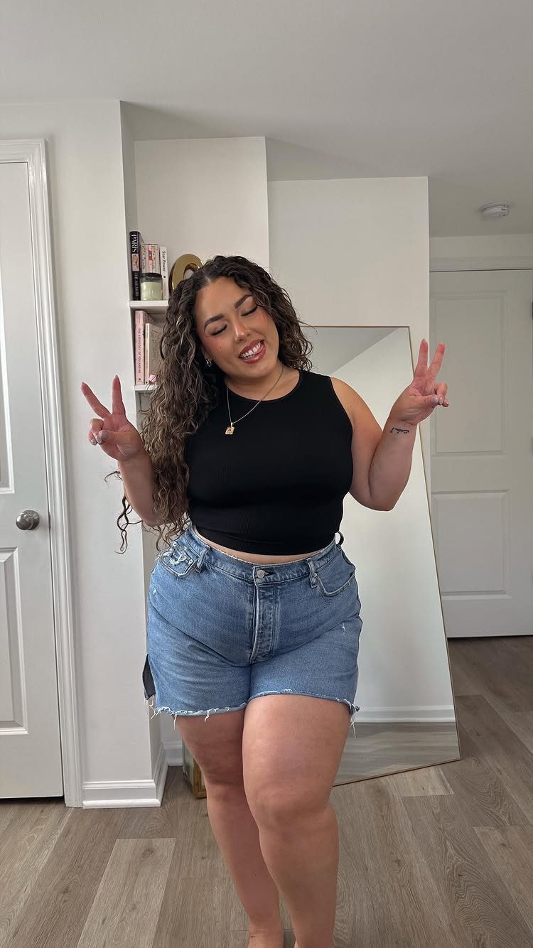 A woman posing in a black crop top and blue denim shorts while making peace signs.