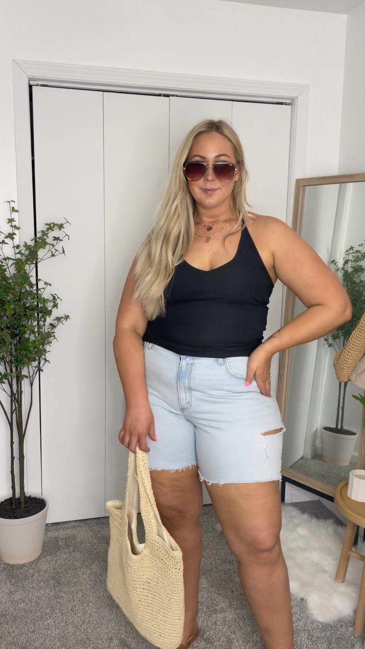 A blonde woman posing in a black tank top and light blue denim shorts.