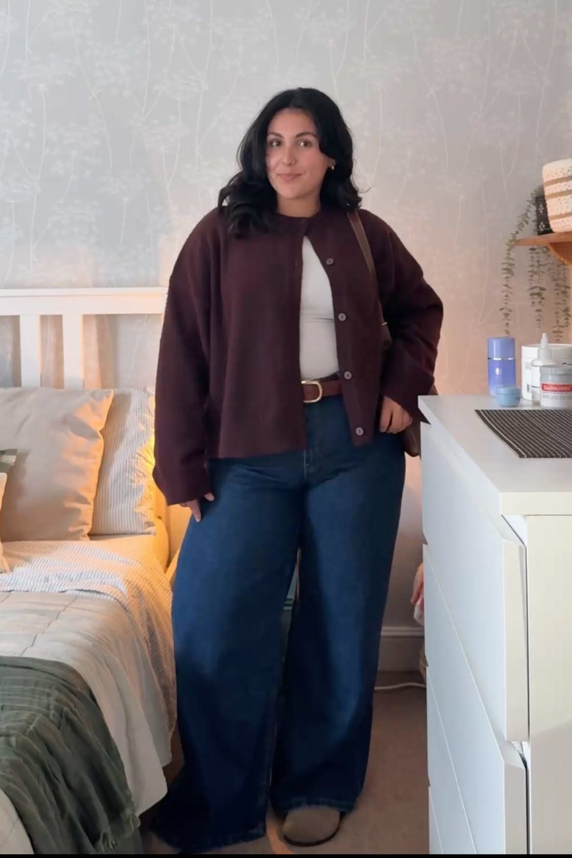 A woman wearing a burgundy knit cardigan, white top, and blue wide-leg jeans.