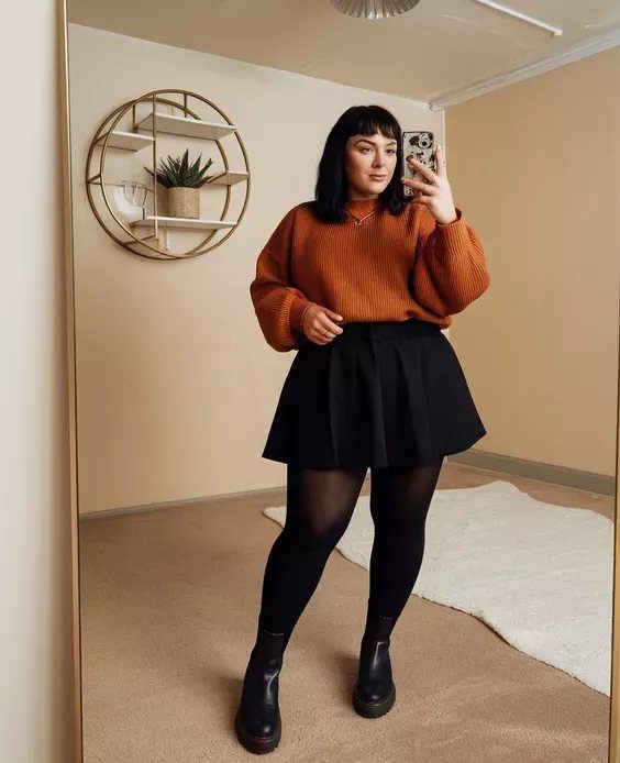 A woman posing in a rust sweater, black skirt, and black tights for a fall outfit.