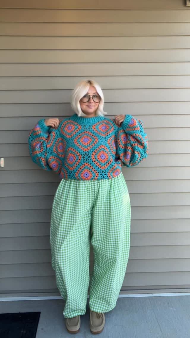 Blonde woman posing in a multicolored crocheted cropped sweater and green checkered trousers.