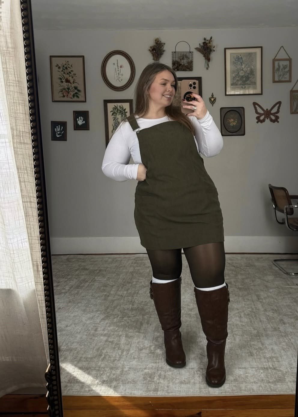 Woman posing in a mirror wearing an olive green pinafore dress, white shirt, and brown boots.