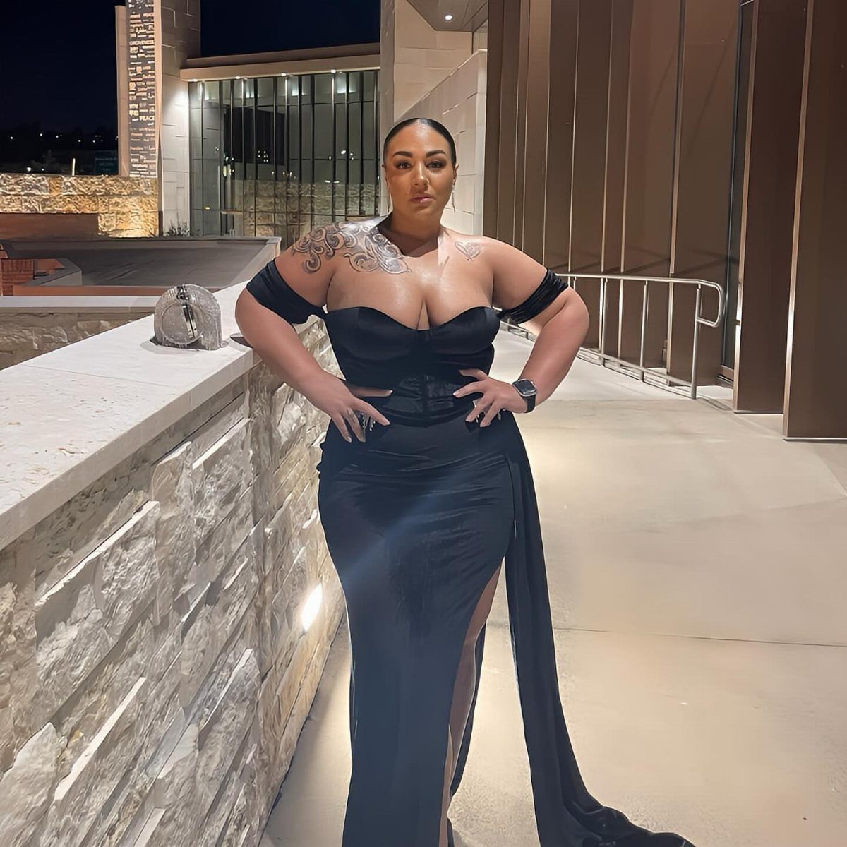 A plus size woman posing in a black off-the-shoulder evening gown outdoors at night.