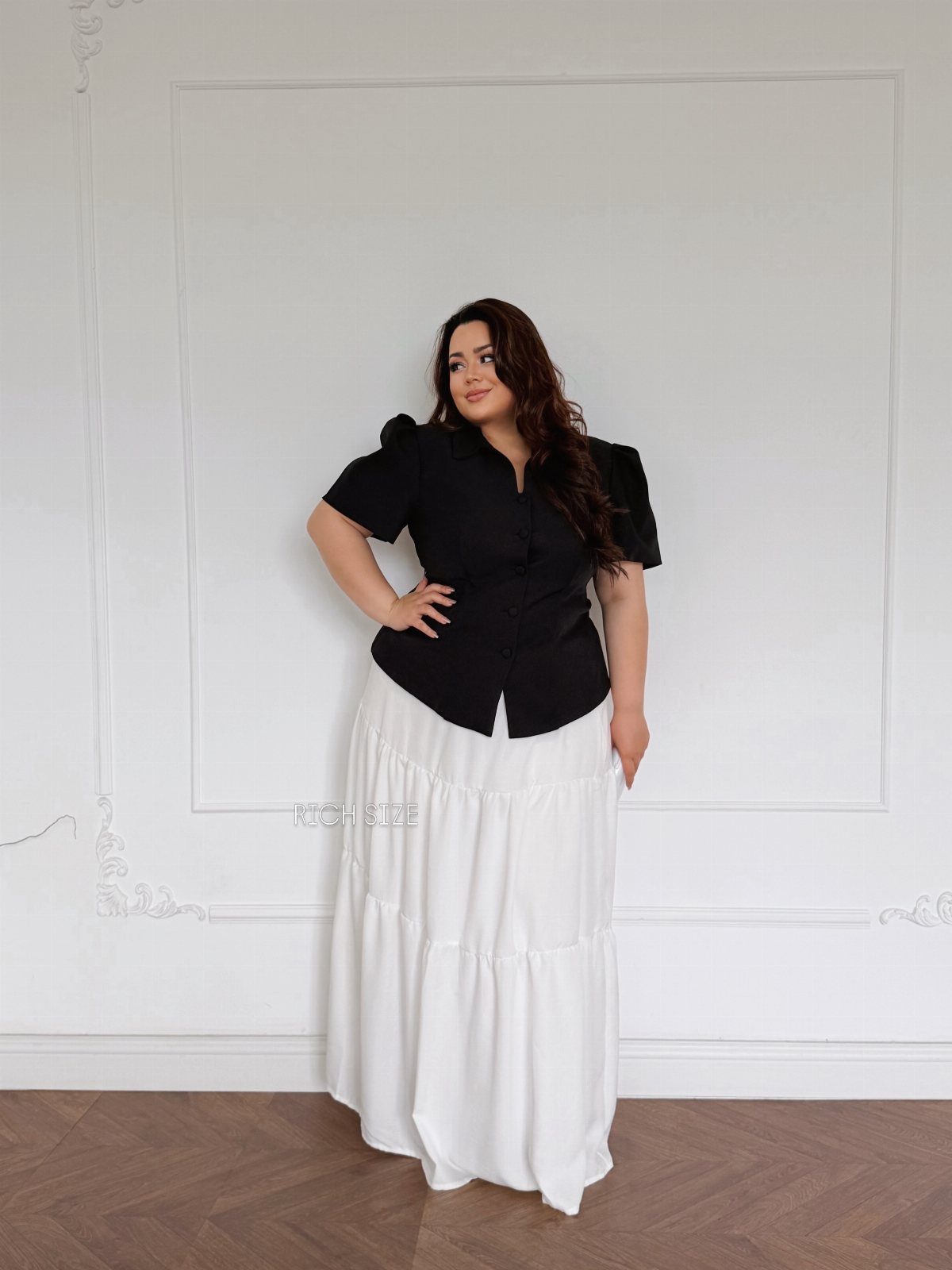 A plus size woman posing in a black blouse and long white tiered skirt.