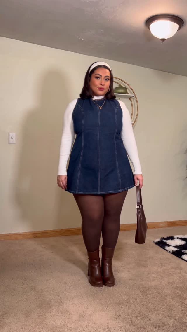 A woman wearing a denim mini dress, white turtleneck, dark tights, and brown boots.