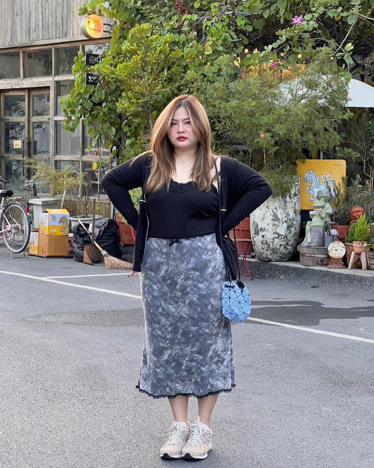 A plus size woman posing in a black top and grey patterned midi skirt outdoors.
