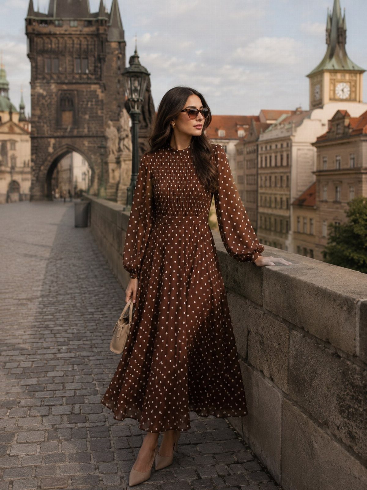 Woman in a brown polka dot midi dress standing on a cobblestone bridge in a city.